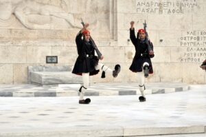 two men in uniform holding guns with Syntagma Square in the background